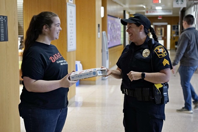 A smiling police officer hands a young lady a pizza.